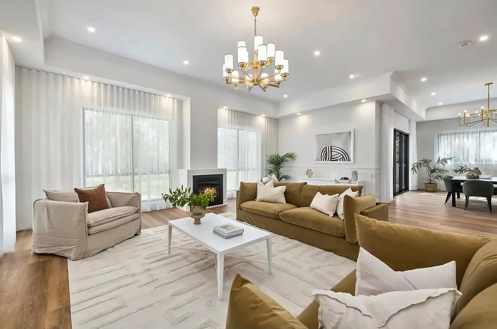 A formal lounge with a chandelier, a fireplace, sheer curtains and soft ochre sofas
