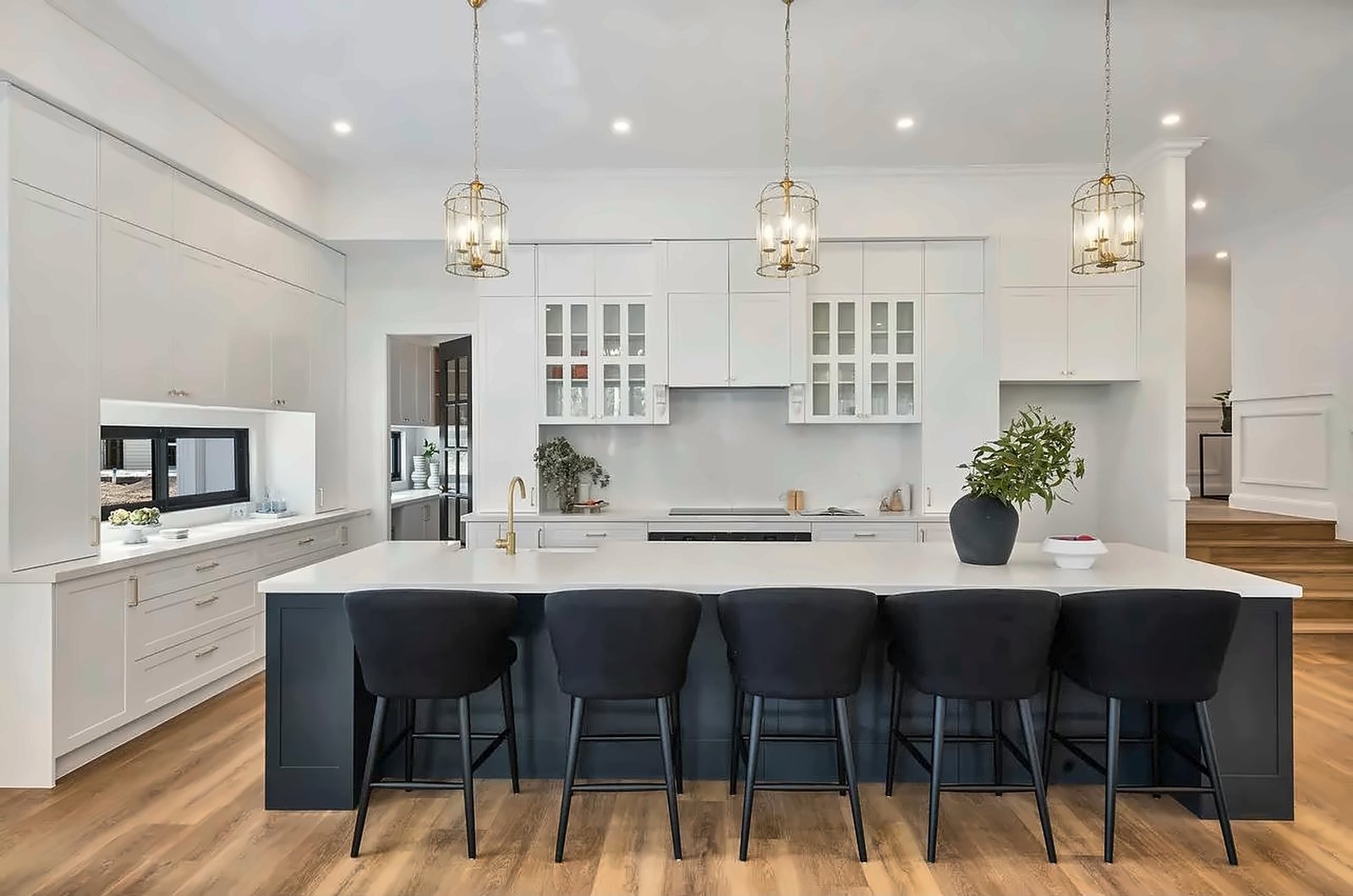 A white Hamptons kitchen with glass-front cabinetry, brass pendant lights and a long charcoal island with stools